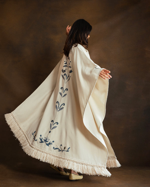 A woman wearing a flowing cream-colored kaftan with blue floral handembroidery and fringe trim, captured from behind as she twirls against a dark brown studio backdrop.