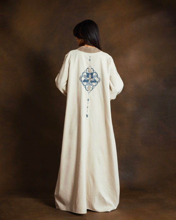 Back view of the YMZ Mosaico Open Kaftan in cream wool blend, featuring intricate blue mosaic-inspired hand embroidery across the back, photographed against a dark brown studio backdrop.