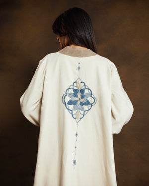 Back view of the YMZ Mosaico Open Kaftan in cream wool blend, featuring intricate blue mosaic-inspired hand embroidery across the back, photographed against a dark brown studio backdrop.