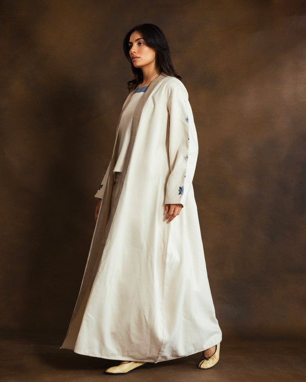Side view of the YMZ Mosaico Open Kaftan in cream wool blend, featuring blue mosaic-inspired hand embroidery on the sleeves and a relaxed, flowing silhouette against a textured brown studio backdrop.